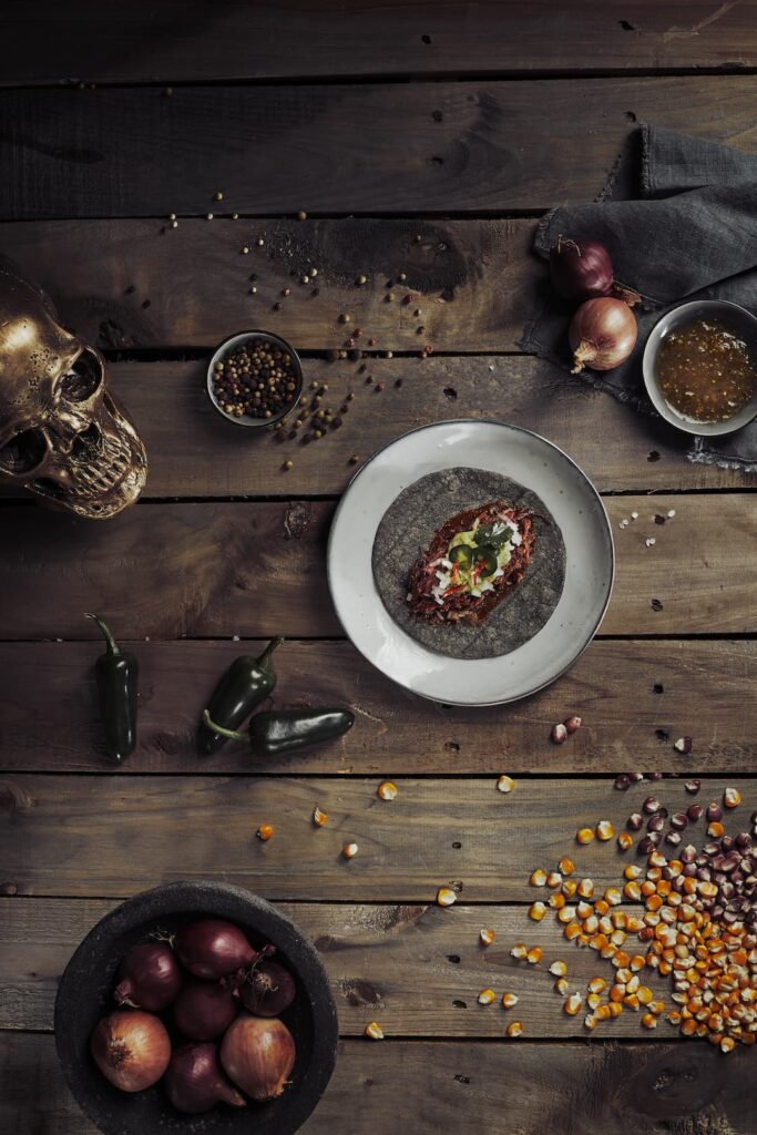 Taco mit Fleisch, Zwiebeln, Koriander und Jalapeño auf einem blauen Tortilla-Fladen, angerichtet auf einem weißen Teller. Drumherum dekoriert mit ganzen Jalapeños, roten Zwiebeln, Pfefferkörnern, einer Schüssel mit Salsa, gelbem und violettem Mais, sowie einem dekorativen Metallschädel – alles auf rustikalem Holzuntergrund.