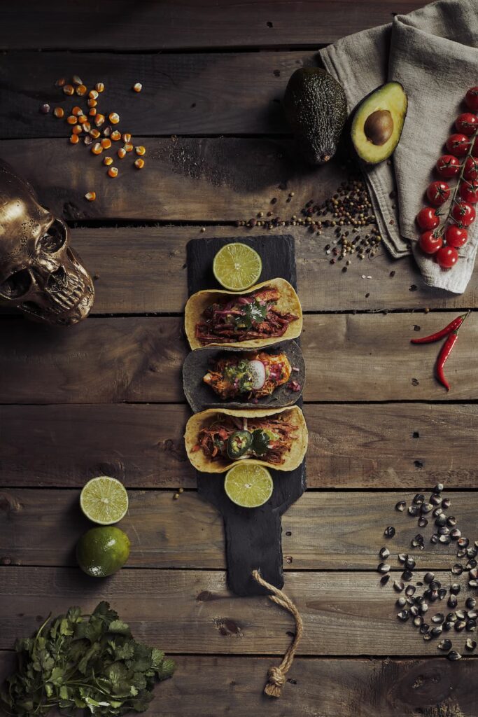 Drei Tacos auf einem schwarzen Servierbrett mit Limettenscheiben, angerichtet auf rustikalem Holztisch. Die Tacos sind mit Fleisch, roten Zwiebeln, Koriander, Jalapeños und Radieschenscheiben gefüllt. Umgeben von einer halbierten Avocado, Kirschtomaten am Zweig, ganzen Limetten, roten Chilischoten, Pfefferkörnern, Maiskörnern, Kräutern und einem dekorativen Metallschädel.