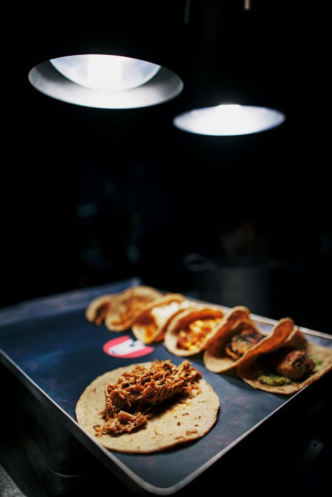 Fünf frisch zubereitete Tacos auf einem dunklen Tablett unter zwei Spotlampen, einzeln belegt mit verschiedenen Füllungen wie mariniertem Fleisch, Guacamole und Käse, stilvoll angerichtet im Holy Taco.