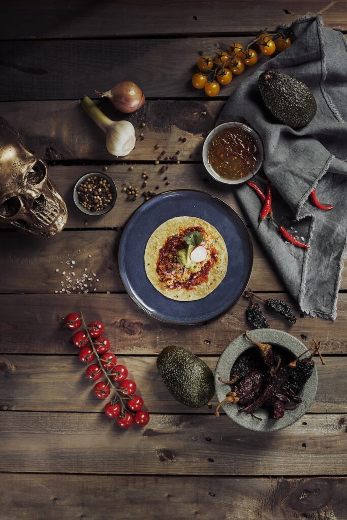 Teller mit einem belegten Taco auf rustikalem Holztisch, umgeben von Knoblauch, Zwiebel, Avocados, roten und gelben Kirschtomaten, Chilischoten, getrockneten Chilis in einer Steinmörserschale, Schale mit Salsa, Pfefferkörnern, Salz und einem goldfarbenen Totenschädel als Dekoration.