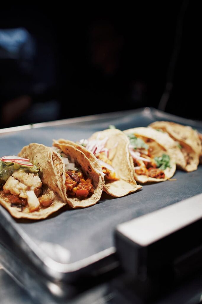 Sechs verschiedene Tacos auf einem Tablett in der Restaurantküche, abwechslungsreich gefüllt mit Gemüse, Fleisch, Salsa, Guacamole und Zwiebeln – stilvoll angerichtet unter hellem Licht.